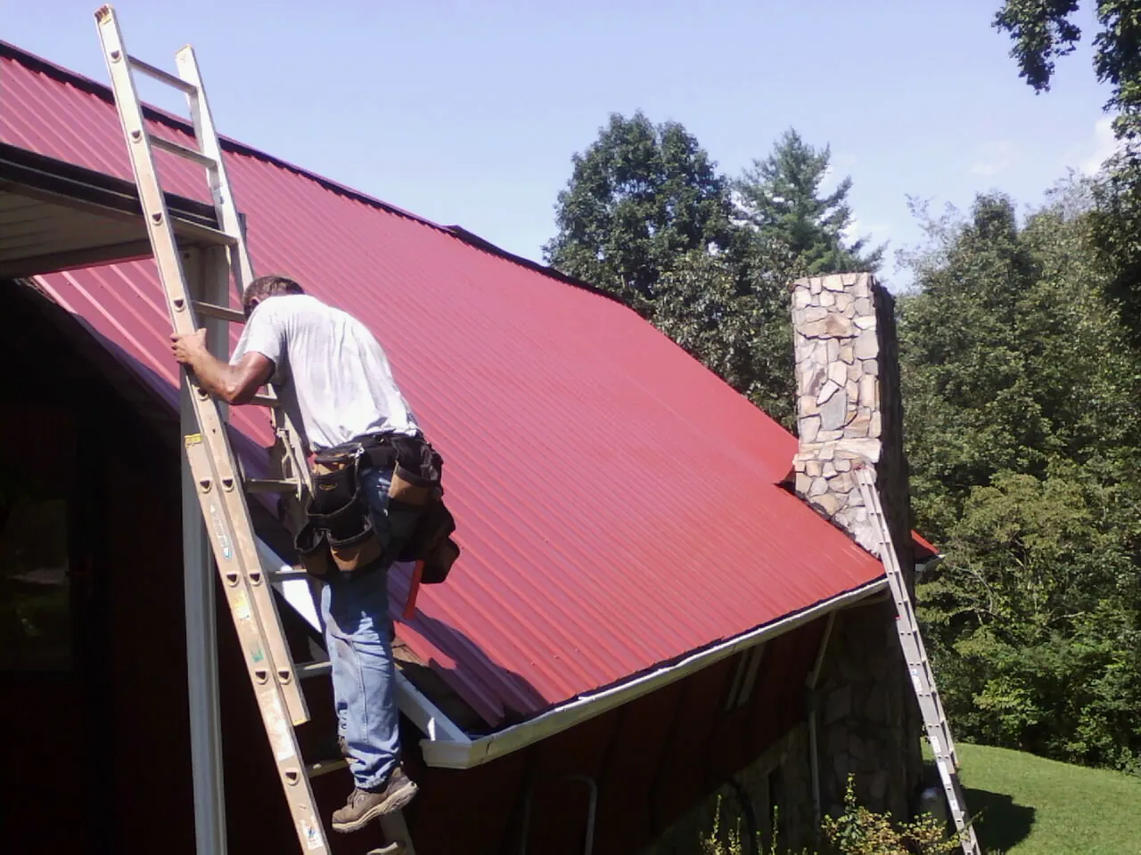 Roofer on a ladder beside a red metal roof and stone chimney — Soffit & Fascia Repair by Roofing Brothers in Ulster Park